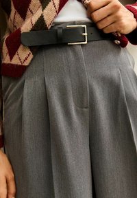 Pantaloni grigi su misura con pince, abbinati a una cintura in pelle nera con fibbia dorata. Tessuto texturizzato con un motivo sottile.