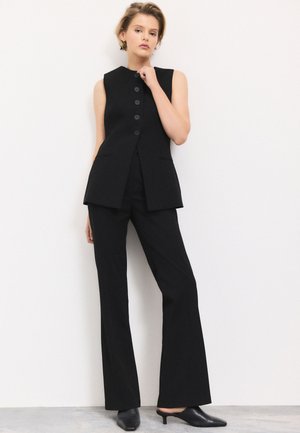 Frau in ärmelloser schwarzer, geknöpfter Weste und weitem Hosenbein, trägt schwarze mules mit Absatz, steht vor weißem Hintergrund.