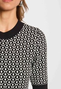 Frau trägt ein kurzärmliges Top mit schwarz-weißer geometrischer Musterung, einem schwarzen gerippten Kragen und silbernen Creolen.