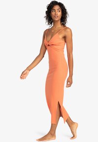 Oranje aansluitende jurk met spaghettibandjes, knoopdetail bij de buste en hoge splitten tot de dij. Zachte stof met een gladde textuur.