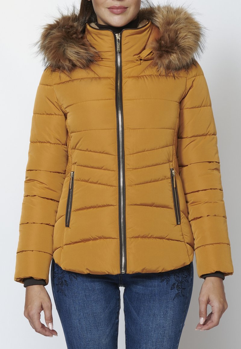 Femme portant une veste matelassée jaune moutarde avec capuche bordée de fourrure et un jean bleu, debout devant un fond blanc uni.