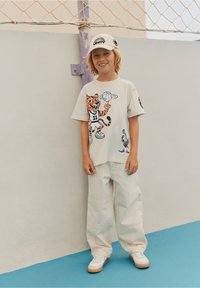 Niño sonriendo con una gorra blanca, camiseta grande con un gráfico de tigre, sosteniendo un baloncesto giratorio, pantalones amplios claros y zapatillas blancas sobre un suelo azul.