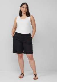Schwarze Shorts mit lockerer Passform, elastischem Bund und Kordelzug. Kombiniert mit einem weißen Tanktop und braunen flachen Sandalen. Einheitlicher Hintergrund.