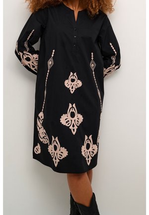 Mujer con un vestido negro hasta la rodilla con escote abotonado y bordado floral beige en el frente y las mangas, combinado con botas negras.