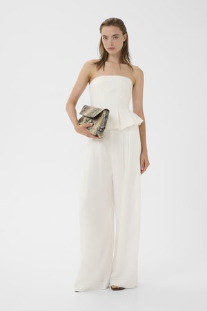 Frau trägt ein trägerloses weißes Peplum-Top und weiße, weit geschnittene Hosen, hält eine große Clutch aus Schlangenleder.