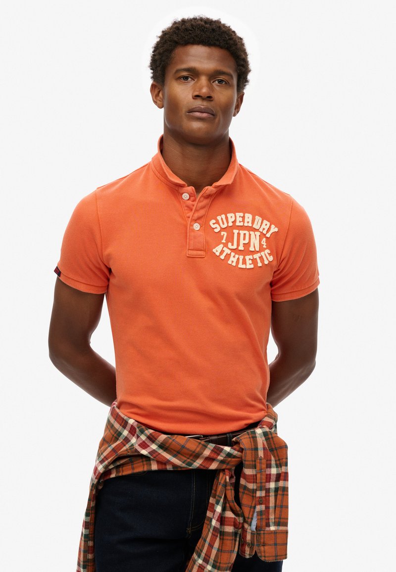 Pomarańczowa koszulka polo z trzema guzikami, z białym napisem „SUPERDRY JPN ATHLETIC”. Noszona z w kratkę pomarańczowo-brązową flanelą zawiązaną w pasie.