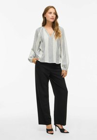Femme se tenant debout avec une main dans la poche, portant un blouse à manches longues blanc et noir à rayures, un pantalon large noir et des talons ouverts noirs.