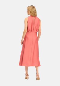 Robe sans manches corail avec un col montant et un dos en forme de trou de serrure. Tissu lisse, longueur mi-mollet, taille élastique et détails de drapés subtils.