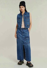 Νεαρή γυναίκα φορώντας ένα αμάνικο denim γιλέκο, μακρά denim φούστα με τσέπες, μαύρο μπερέ, μπεζ μπλούζα και μαύρες πλατφόρμες.