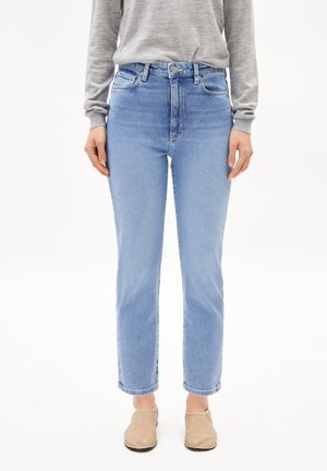 Personne portant un jean droit taille haute bleu clair, un haut gris à manches longues et des chaussures béiges à enfiler, debout devant un fond uni.