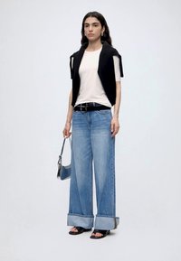 Femme portant un jean large avec revers, ceinture noire, t-shirt blanc, pull noir porté sur les épaules, sandales noires, tenant un petit sac à main.