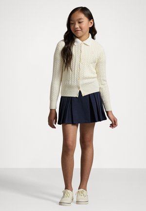 Cremfarbener Strickcardigan mit Knöpfen, kombiniert mit einem marineblauen Faltenrock und hellen Sneakers. Einfaches Design mit strukturiertem Schnitt.