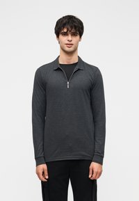 Σκούρο γκρι μπλουζάκι μακρυμάνικο με φερμουάρ στον γιακά, με απαλή υφή και εφαρμοστή σχεδίαση. Φορεμένο πάνω από μαύρο πουκάμισο.