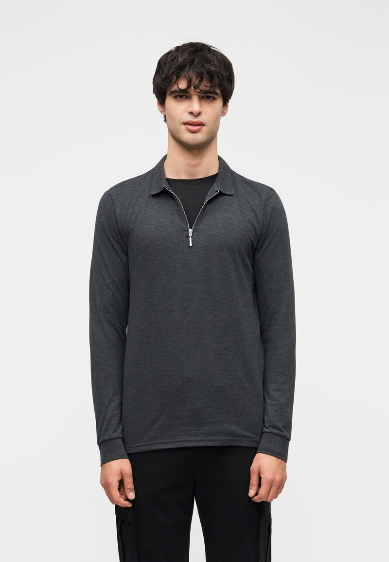 Σκούρο γκρι μπλουζάκι μακρυμάνικο με φερμουάρ στον γιακά, με απαλή υφή και εφαρμοστή σχεδίαση. Φορεμένο πάνω από μαύρο πουκάμισο.