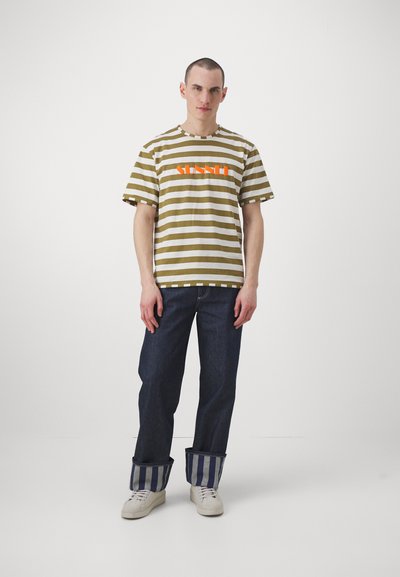 Oliven- og hvit stripet t-skjorte med "SUN" trykket i oransje, sammen med mørkeblå jeans med stripete mansjetter og hvite sneakers.