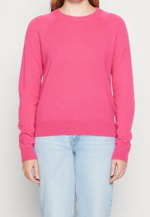 Persona con un suéter de manga larga de color rosa brillante y vaqueros azul claro, de pie frente a un fondo blanco liso.
