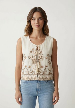 Donna con capelli castani ondulati indossa un top crema senza maniche con ricami floreali marroni e lacci con nappine, abbinato a jeans azzurri chiari.