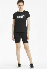 Mujer joven de pie con una camiseta negra de Puma, pantalones cortos de ciclismo negros y zapatillas negras contra un fondo blanco.