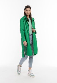 Giacca impermeabile verde con cappuccio, chiusura a zip, tasche laterali e cordino. Abbinata a jeans azzurri chiari e sneaker multicolori.