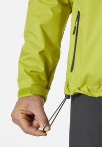 Chaqueta impermeable verde claro hecha de tejido texturizado, con un bolsillo con cremallera y un cordón ajustable en el dobladillo, además de un cierre de muñeca de velcro.