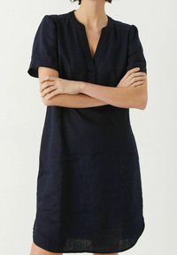 Nainen, jolla on lyhythihainen, tummansininen polvipituinen mekko V-kauluksella, kädet ristissä vartalon edessä yksinkertaista taustaa vasten.