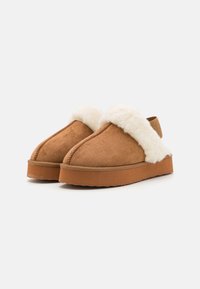 Beige Wildleder-Slipper mit einem flauschigen weißen Fellbesatz. Dicke Gummisohle, abgerundete Zehenform und sichtbare Steppnähte entlang der Kanten.