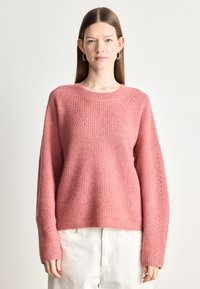 Roza pletena jopica z rebrasto teksturo, okroglo ovratnico in dolgimi rokavi. Ima okrasno šivanje ob šivih. V kompletu z svetlimi hlačami.