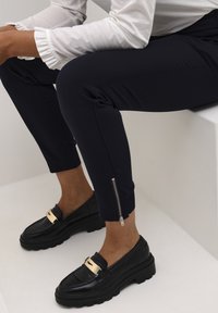 Marinblå kortbyxor med siddragkedjor, kombinerade med svarta loafers med guldaccenter. Slät tygkvalitet med skräddarsydd passform och rena linjer.