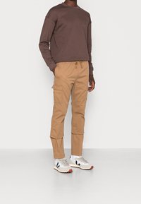 Hnědá mikina s dlouhým rukávem v kombinaci s béžovými cargo kalhotami s bočními kapsami. Boty jsou bílé s námořnickými a červenými detaily.