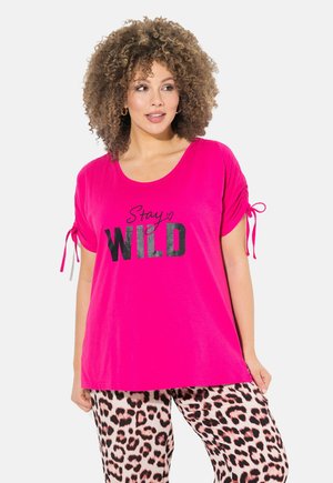 Helles pinkes T-Shirt mit dem Text "Stay WILD" in Schwarz, mit kurzen Ärmeln und gebundenen Akzenten. Kombiniert mit Hosen im Leopardenmuster.