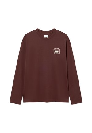 Junge Person mit langen roten Haaren, trägt einen bordeauxfarbenen Pullover und dunkelbraune Hosen, steht mit den Händen in den Taschen und schaut zur Seite.