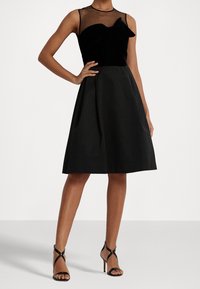 Svart klänning med en sammetstopp med genomskinlig halsringning, en rosettdetalj och en utsvängd kjol av tyg. I kombination med svarta sandaler med remmar.