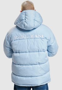 Lyseblå dunjakke med hette. Har horisontale quiltede seksjoner og brodert "KARL KANI"-logo på ryggen. Glatt tekstur.