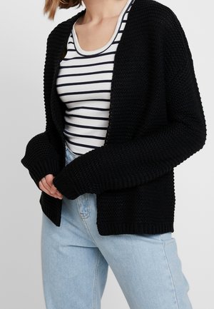 Frau trägt eine schwarze Strickjacke über einem weiß-schwarz gestreiften Tanktop, kombiniert mit hellblauen Jeans.