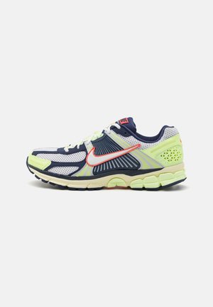 Chaussure de sport basse blanche, bleu marine et jaune néon avec un Swoosh Nike rouge et une semelle amortie, vue de côté sur un fond blanc.