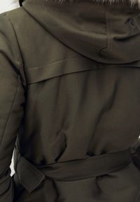 Oliwkowa parka z kapturem wykończonym futrem, z paskiem w talii i gładką teksturą tkaniny; posiada projekt z tylnym yoke oraz subtelne detale szycia.