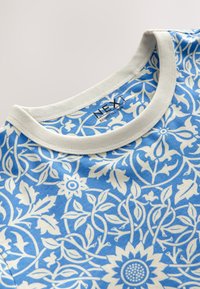 Blaues T-Shirt mit floralem Muster und weißem Kragen. Verziert mit organischen Formen und filigranen Designs auf weichem Stoff. Markenetikett am Kragen sichtbar.