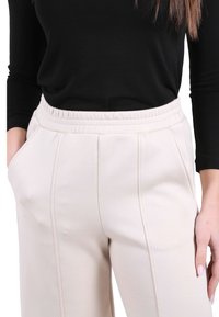 Donna che indossa pantaloni beige a vita alta con pieghe frontali e tasche laterali, abbinati a una maglia nera a maniche lunghe, in piedi con le mani vicino alle tasche.