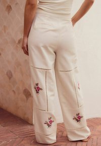 Pantaloni ampi color crema con ricami floreali, dotati di dettagli a intaglio lungo le cuciture e una tasca posteriore. Tessuto morbido.