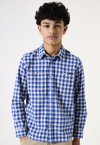 Camisa de cuadros azul y blanca con botones y mangas largas, dos bolsillos en el pecho y una etiqueta de logo. La tela parece tener textura, adecuada para un uso casual.