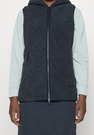 Persoon draagt een zwart fleecevest met ritssluiting over een lichtblauw shirt met lange mouwen en een zwarte doorgestikte rok, staand met ontspannen handen.