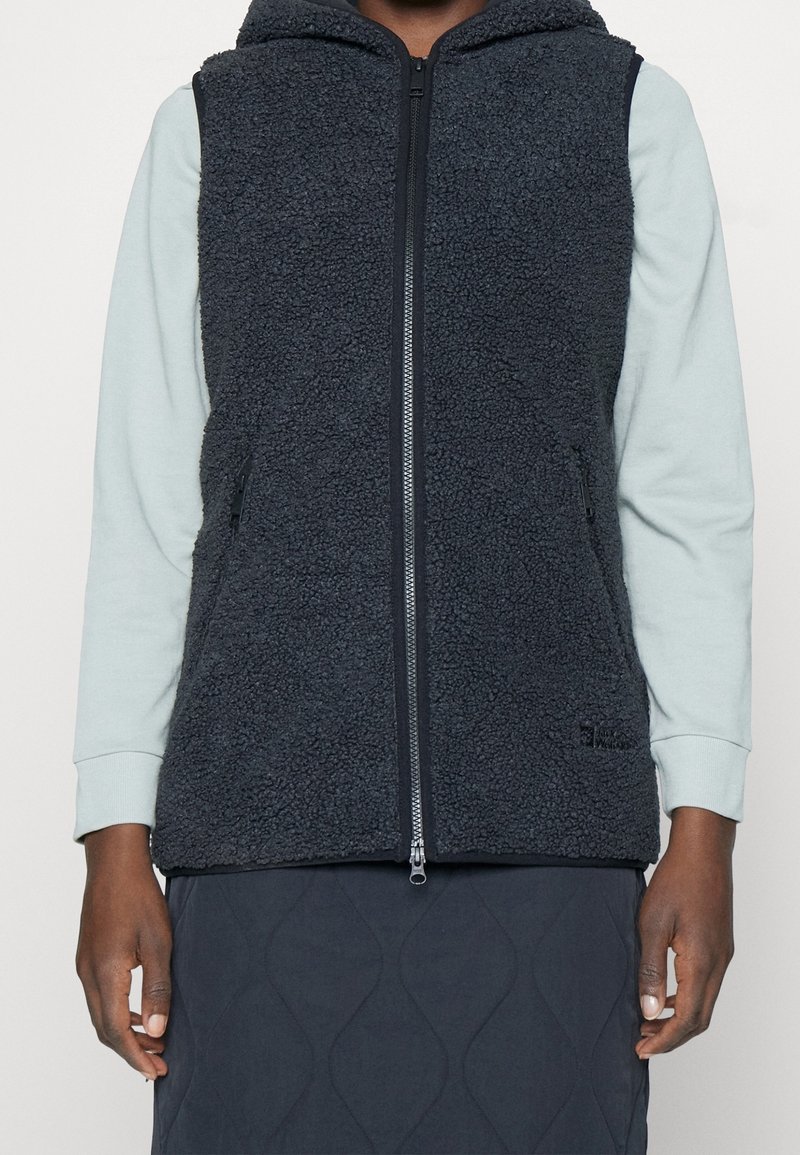 Personne portant une veste polaire noire zippée sur une chemise bleu clair à manches longues et une jupe noire matelassée, debout les mains détendues.