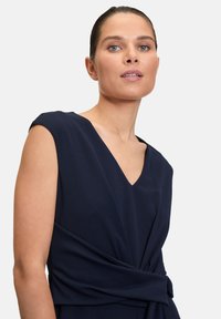 Robe bleu marine avec de courtes manches capes, un décolleté en V et une taille drapée. Texture lisse sans motifs ni détails métalliques.