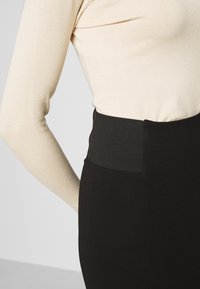 Jupe noire avec une large ceinture noire texturée, associée à un haut ajusté à manches longues de couleur beige clair. Tissu lisse et design épuré.