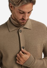 Beige stickad cardigan med krage, två knappar och ribbad manschett. Texturerat mönster ger djup åt tyget.