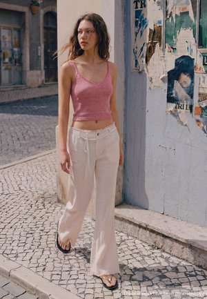 Mladá žena s rozčesanými vlasmi, oblečená v ružovom tielku bez rukávov, bielych širokých nohaviciach a žabkách, kráča po dláždenej ulici.