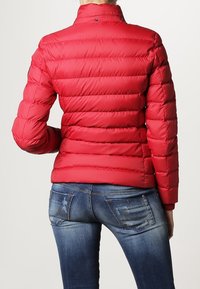 Rød pufferjakke med horisontalt quiltede sektioner, høj krave og trykknaplukning. Sammen med blå denim jeans med baglommer.