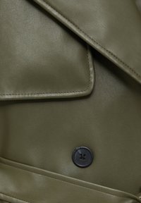 Gros plan d'un manteau en cuir vert olive avec un bouton noir à quatre trous et un détail de poche à rabat cousue.