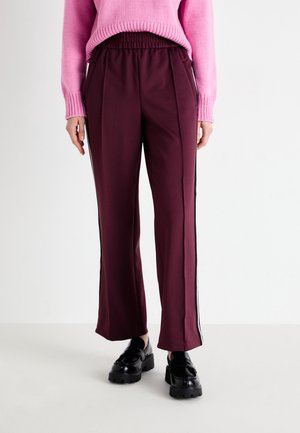 Osoba ubrana w bordowe spodnie o szerokich nogawkach z białymi paskami po bokach, różowy sweter i czarne masywne mokasyny na białym tle.