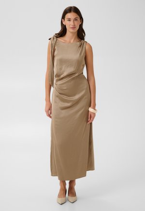 Femme portant une robe sans manches en satin beige avec un détail de nœud sur le côté, des talons pointus beiges et un bracelet blanc, debout sur un fond uni.